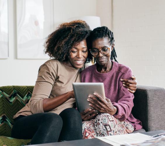 A younger woman sits with her arm around an older woman wearing glasses while they both interact with a tablet.