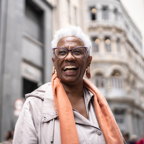 Woman with short white hair, glasses and peach colored scarf smiling outside with city buildings in the background.