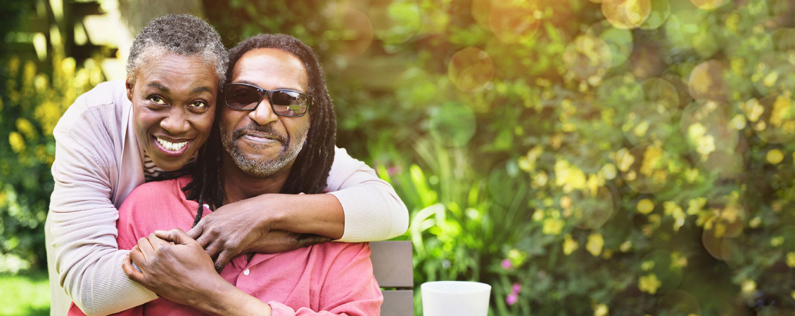 A woman with her arms wrapped around a man wearing dark sunglasses, sitting in a garden.